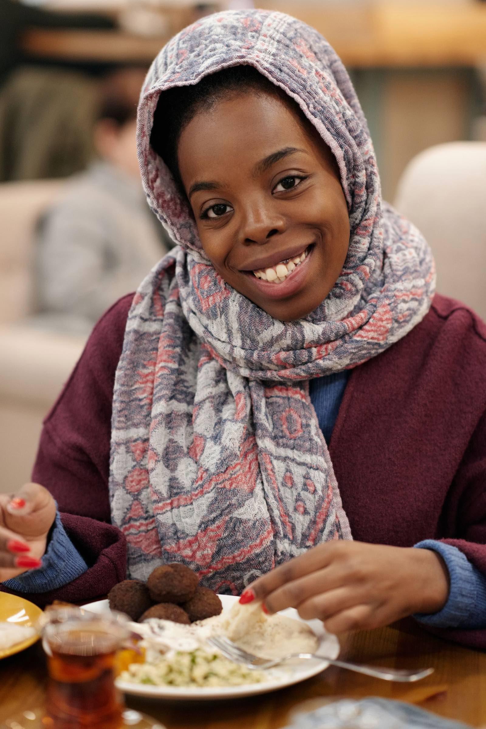A smiling woman in a patterned headscarf at a restaurant table, sharing a plate of falafel and salad with a glass of tea