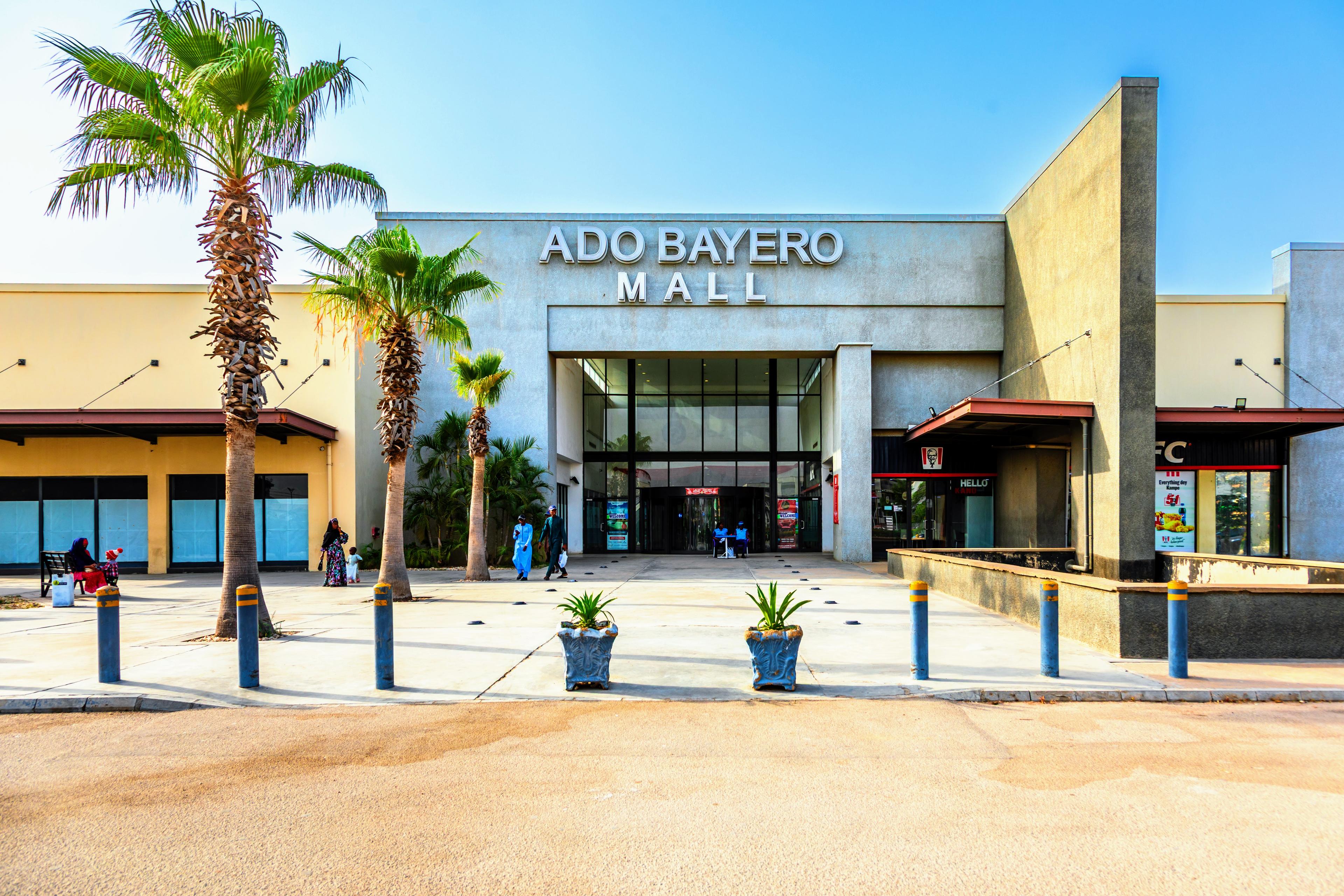 The front entrance of Ado Bayero Mall, with palm trees, visitors walking in, and KFC signage